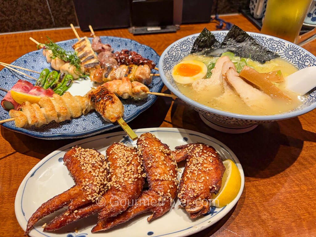 A meal served on a table includes a plate of skewers, a plate of chicken wings, and a bowl of ramen.