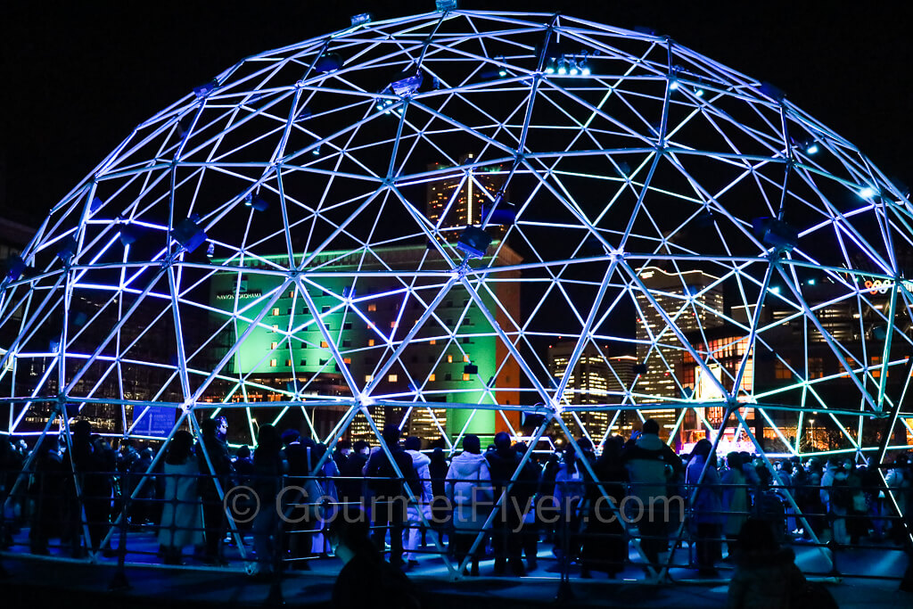 A large dome built with light tubes is displayed in purple and blue lights. Many people are inside the dome.