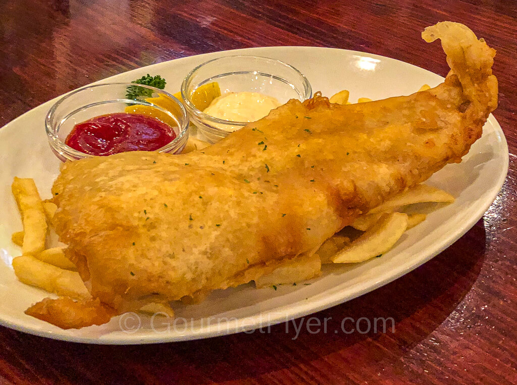 A large piece of British style fried fish is served with French fries and accompanied by small containers of ketchup and tartar sauce.