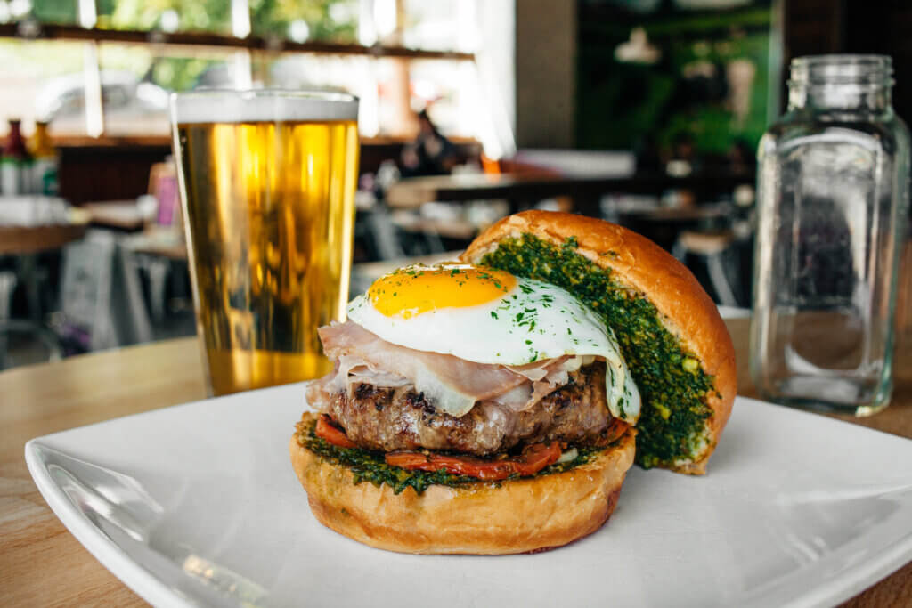 A burger is topped with mozzarella, prosciutto, a sunny side up egg and a   layer of basil pesto.