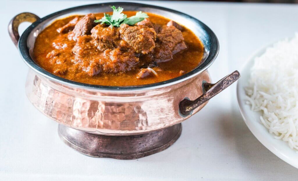 Rich lamb curry with lots of sauce is served in a traditional Indian silver serving bowl.