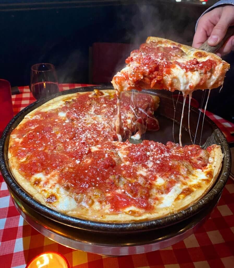 A slice of a deep-dish pizza in its metal dish with a rich tomato topping is served by a spatula.