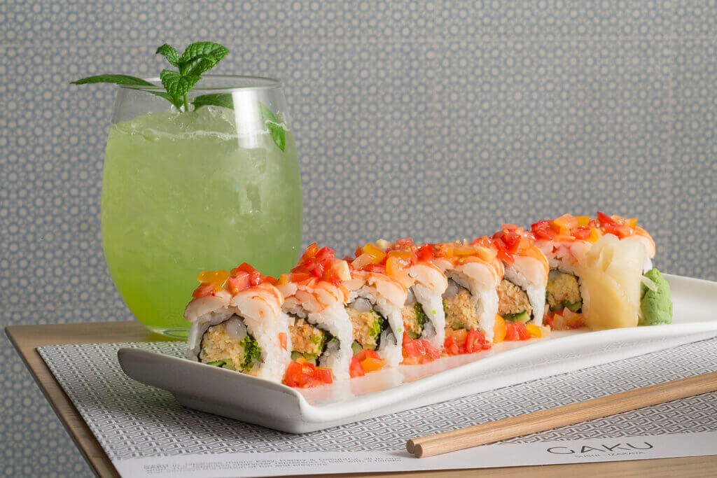 A long platter of sushi cut rolls garnished with crab meat on top is paired with a glass of green beverage on ice with a mint leaf in the background.