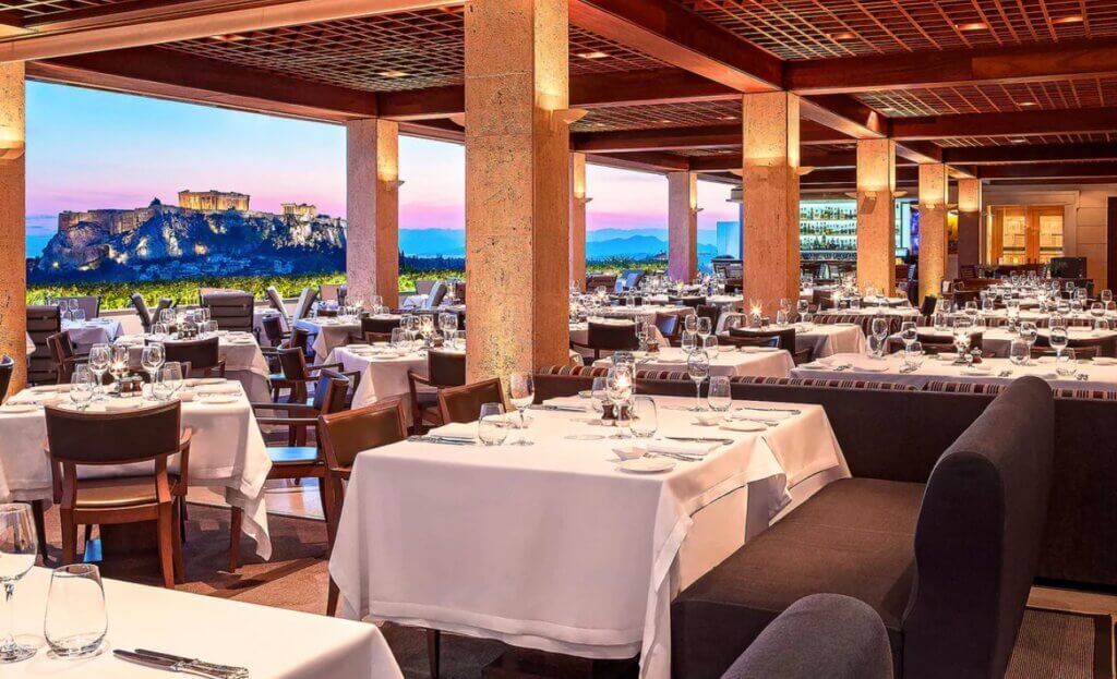 A roof top restaurant with an array of tables with white tablecloth and dinner settings. The Acropolis can be seen in the background.