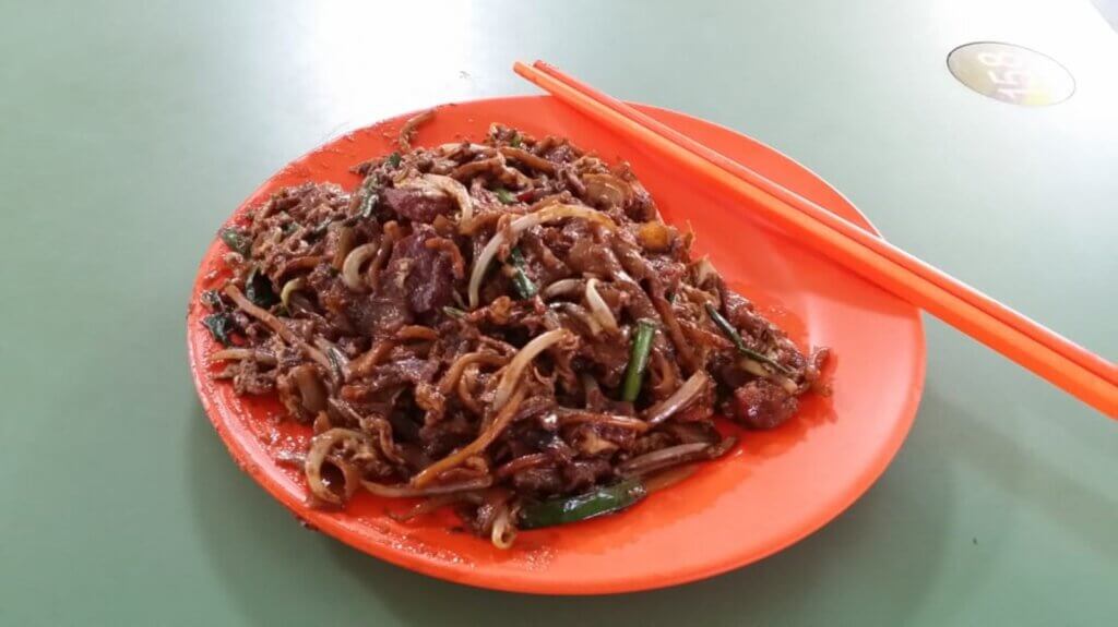 Stir-fried rice noodles topped with meat and bean sprouts is served on an orange plate with orange chopsticks.