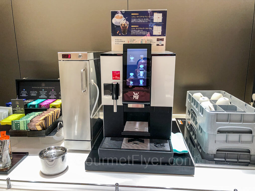 An espresso machine is accompanied by a rack of teabags to the left and trays of cups on the right.