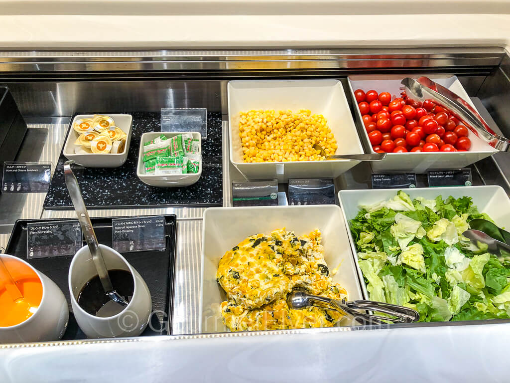 A salad bar with lettuce, corn, cherry tomatoes, pumpkin salad, and two serving bowls of dressings.