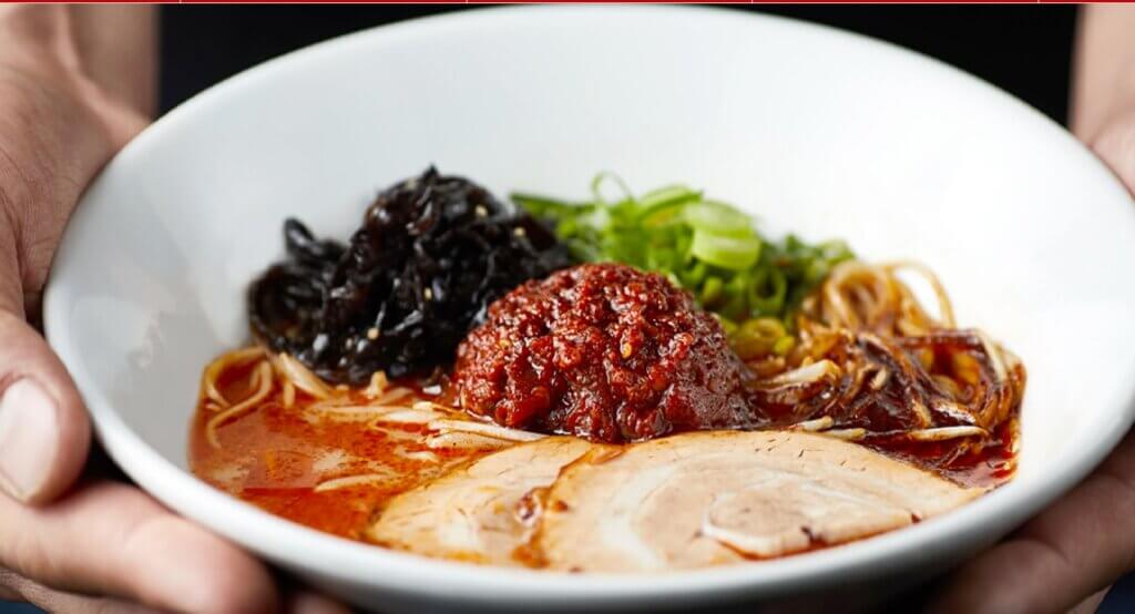 A bowl of ramen is topped with chashu, green onions, other veggies, and chili paste.