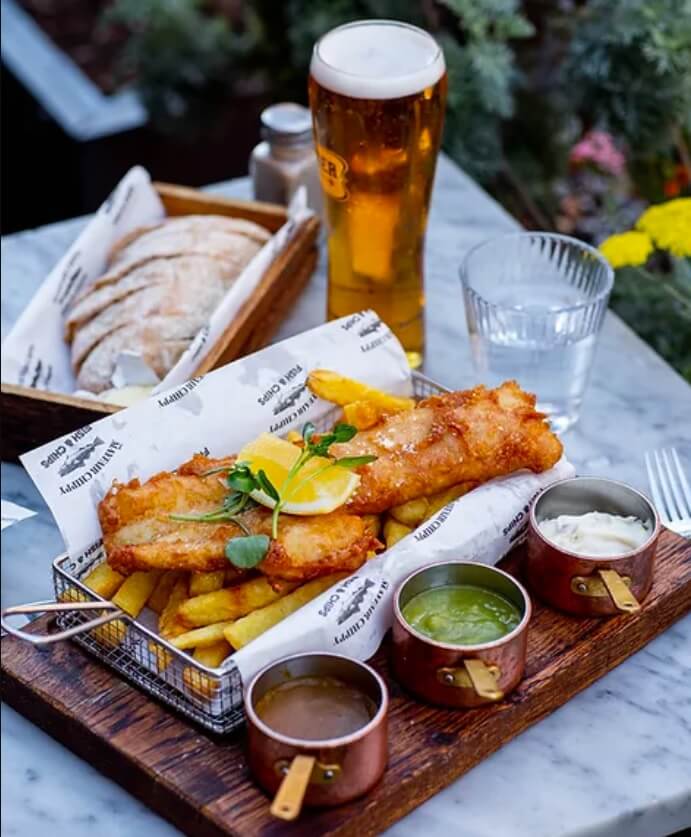 A basket of fish and chips is accompanied by sauces on the side, a loaf of bread, and a glass of beer.