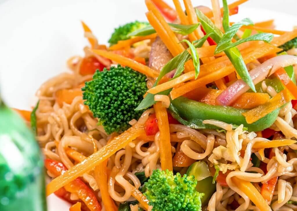 A plate of Asian stir-fried noodles topped with a blend of vegetables including broccoli, shredded carrots and green beans.
