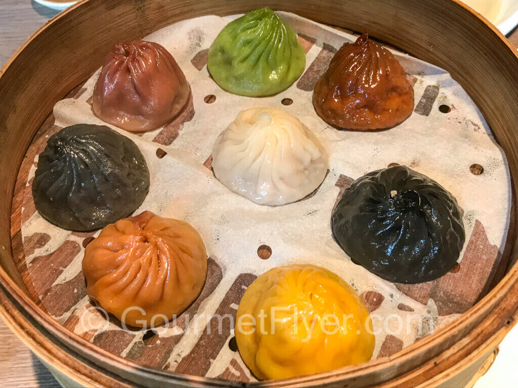 A steamer with a white dumpling in the center is surrounded by 7 other dumplings of various colors.