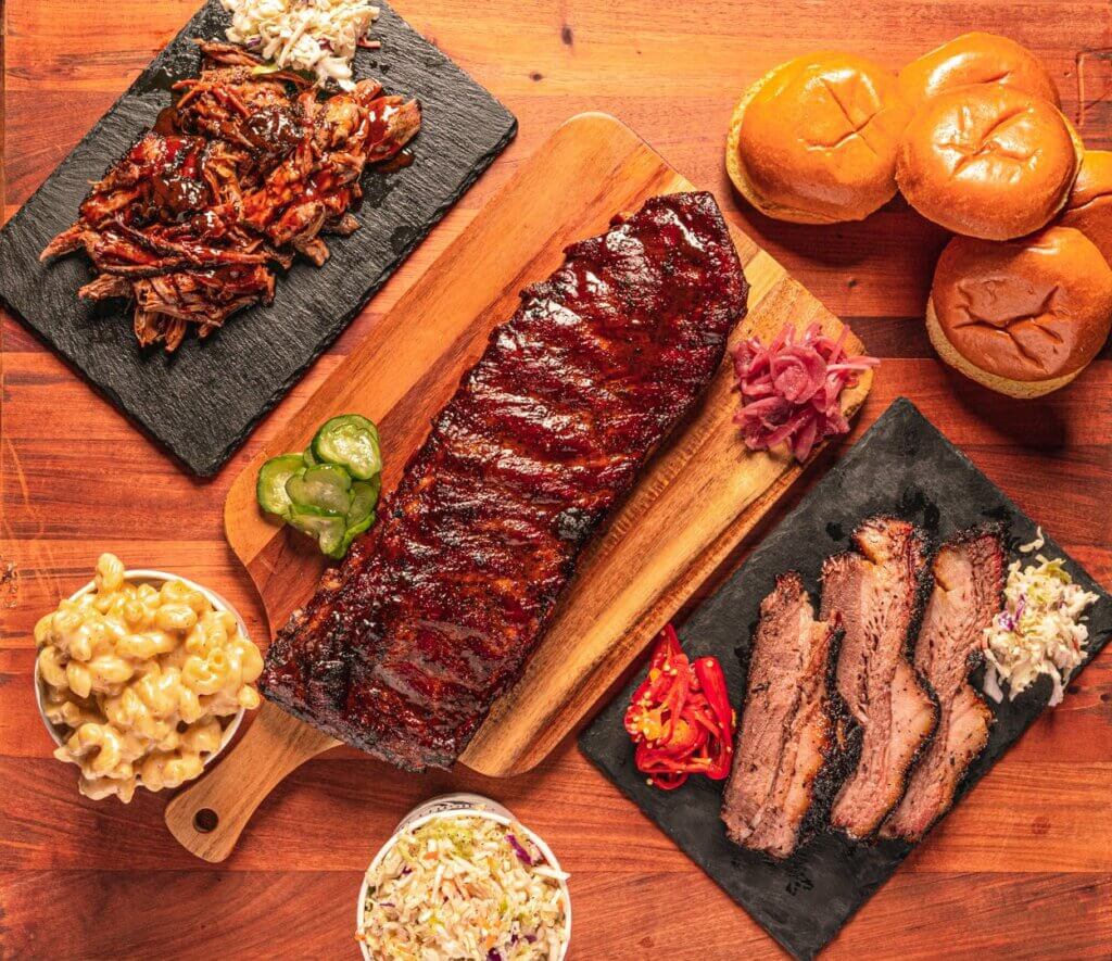 A barbeque platter with a rack of spareribs, a board of sliced brisket and a board of pulled pork are accompanied by buns and macaroni sides.