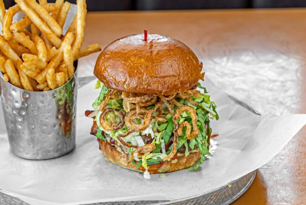 A burger stacked with shredded lettuce and fried onion strings is accompanied by a serving of French fries in a silver cylindrical container.