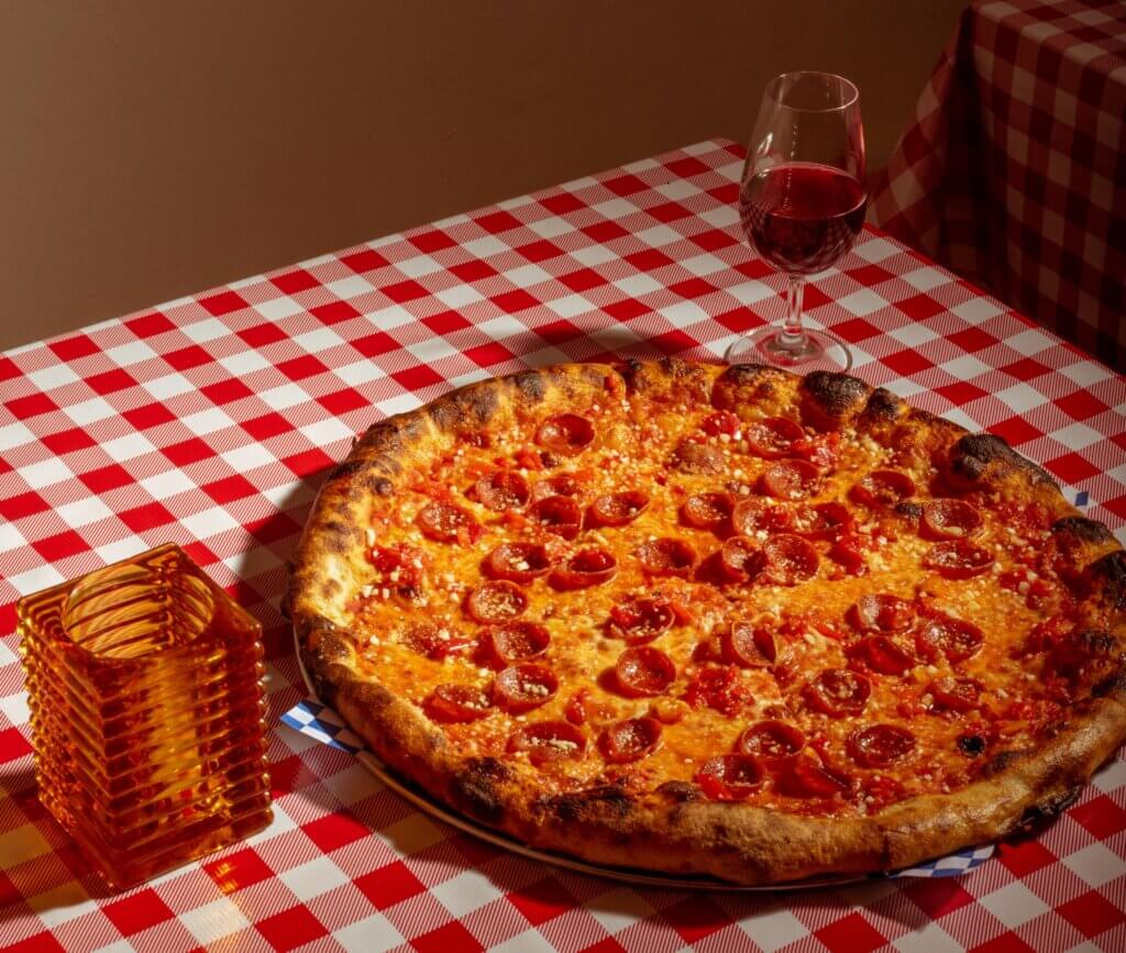 A large pizza with slightly burnt crust is topped with pepperoni and accompanied by half a glass of red wine.