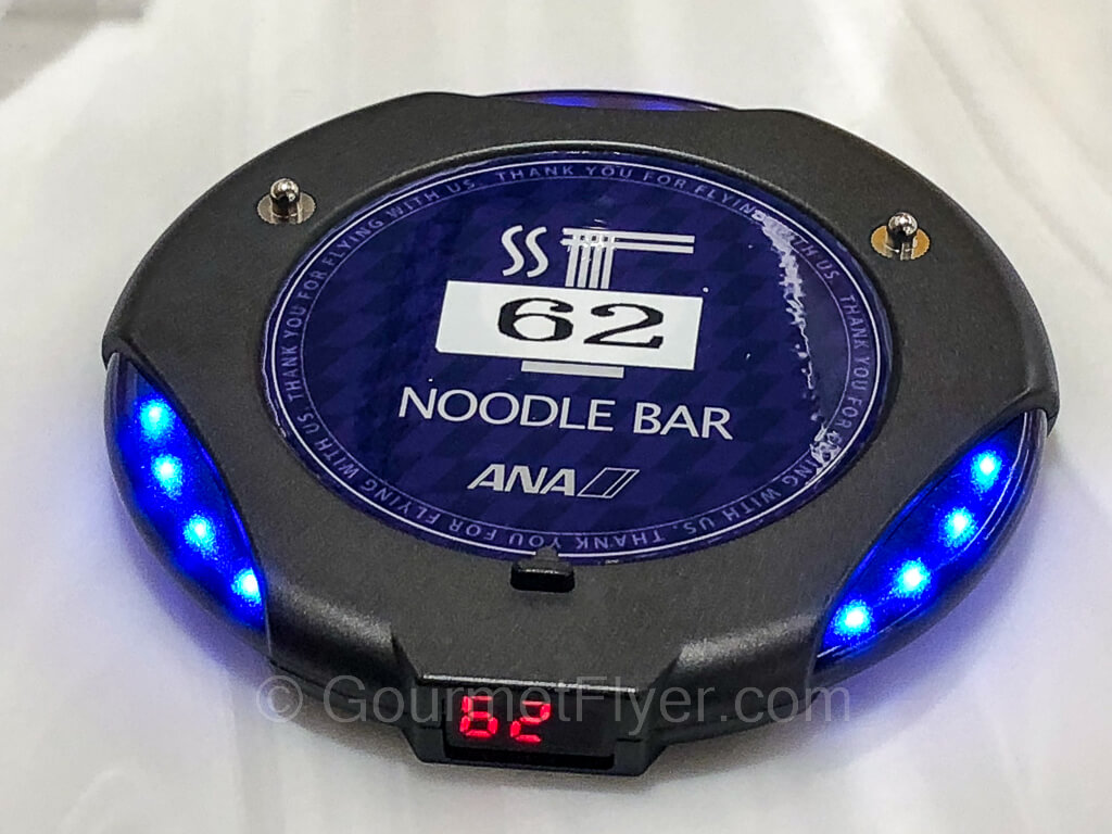 A restaurant style buzzer with the ANA Noodle Bar graphics has its blue LED lights lit up.