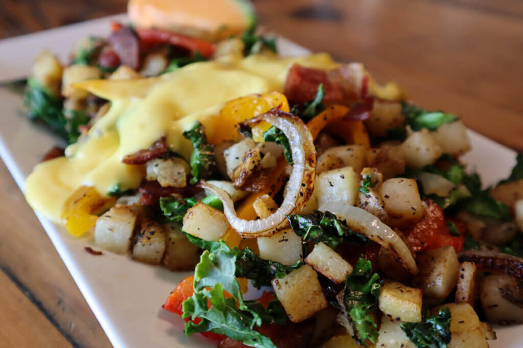 A breakfast platter with sauteed diced potatoes mixed with onions and peppers. An egg Benedict is blurred in the background.