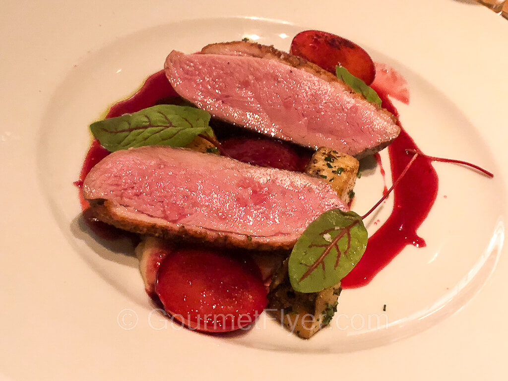 Two slices of medium rare duck breast are served on a plate and garnished with a red fruity sauce.