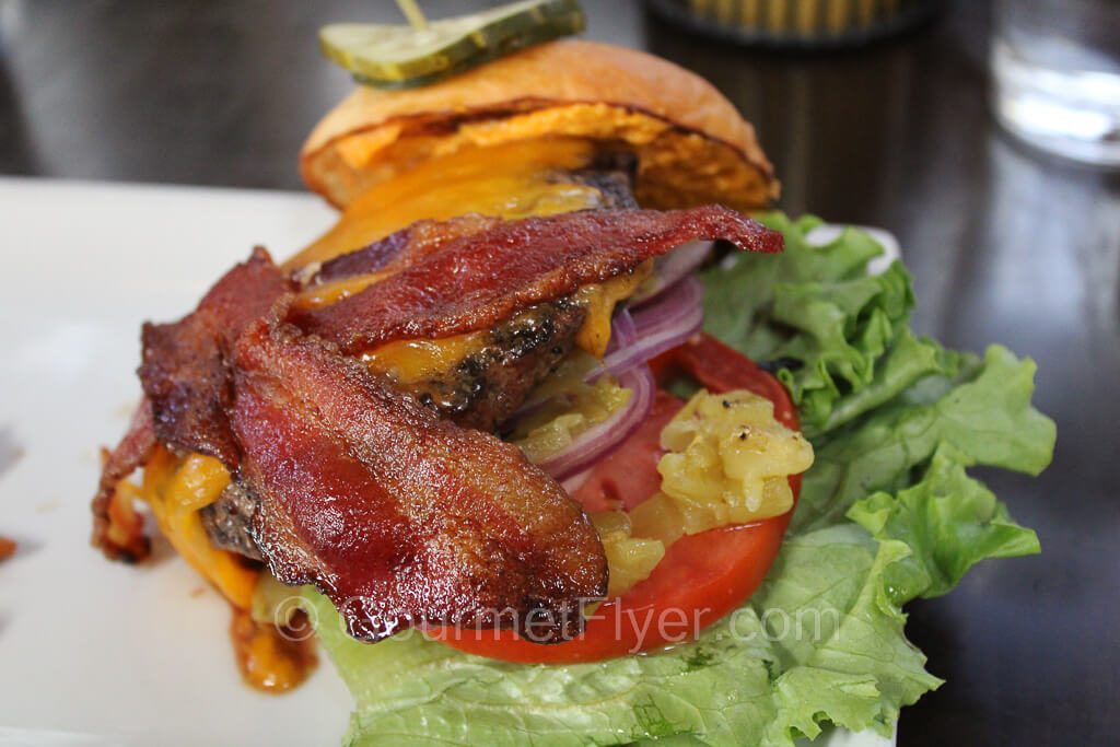 A burger is loaded with cheese, bacon, and green chili which sit atop a beef patty, lettuce, and a slice of tomato.