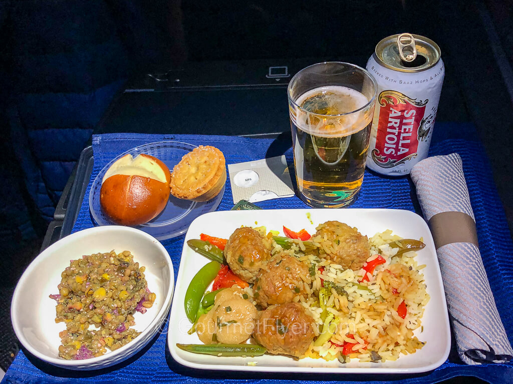 A dinner tray with a dish of meatballs and a side of salad is served with a roll, a small pie, and a beer.