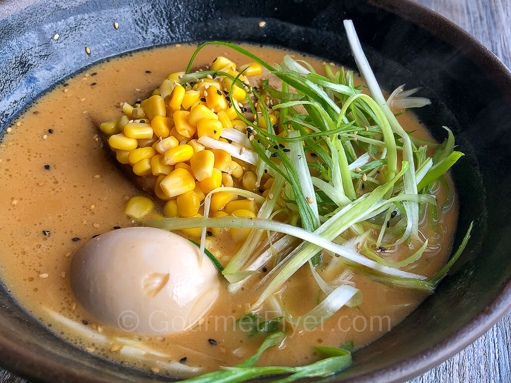 A bowl of ramen topped with meat, an egg, corn, and green onions.