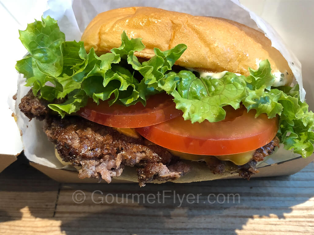 A burger is topped with a large leaf of lettuce and two slices of tomatoes and wrapped in a white paper pocket.