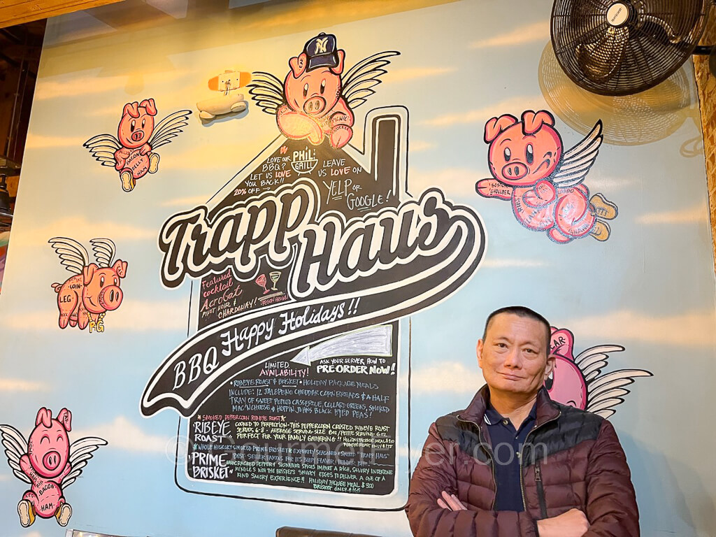 The Gourmet Flyer is posing in front of the wall with the Trapp Haus logo and a half dozen laughing flying pigs.