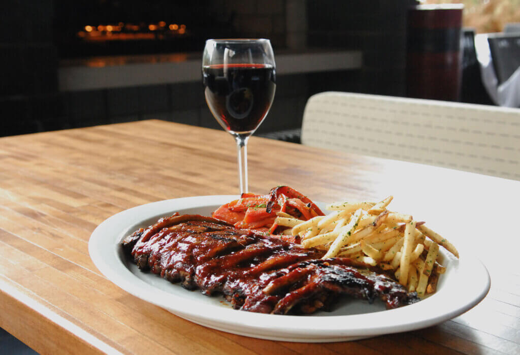 A rack of BBQ ribs is served on a plate with skinny French fries on the side. A glass of red wine sits in the background.