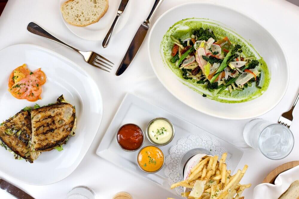 A plate of sandwiches, pasta and a side of French fries are served on a table with white tablecloth and silverware.