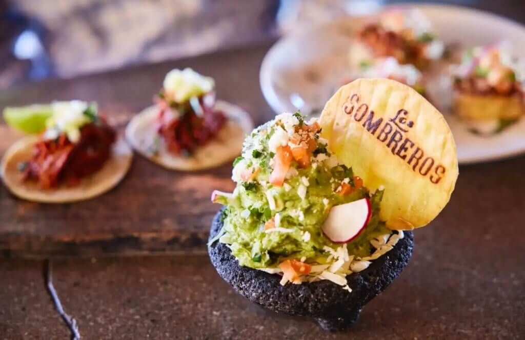 A small taco loaded with guacamole and other garnishes is served with a chip with the "Los Sombreros" logo.