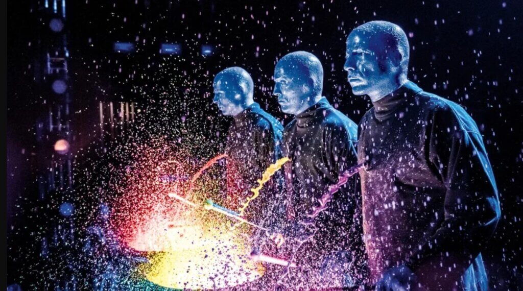 Drums with paint are beaten violently by three blue men, sending neon droplets flying off the stage.