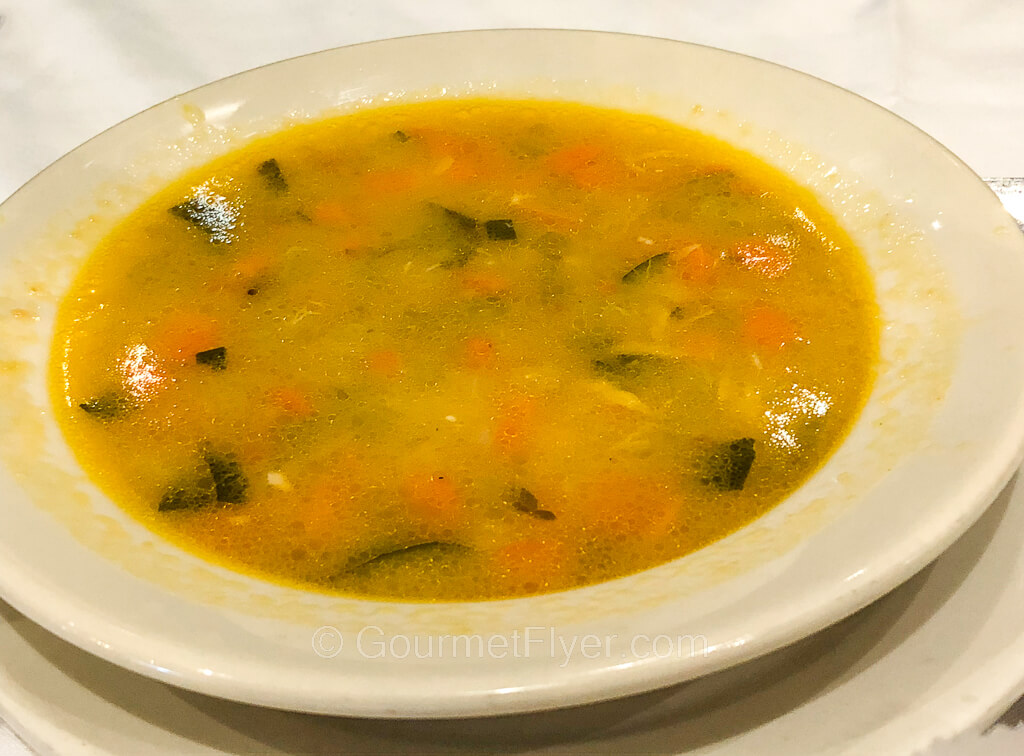A bowl of clear orange soup with carrots, veggies, and small pieces of fish.