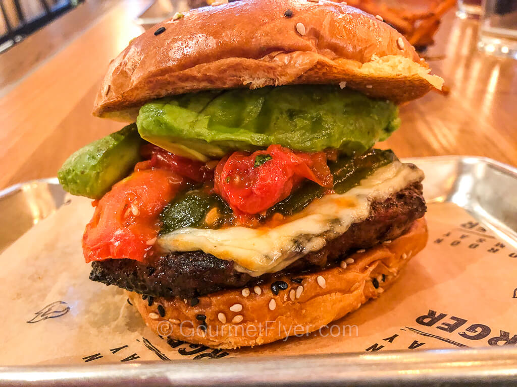 A burger topped with avocado, mozzarella, roasted jalapeño, and roasted tomato.