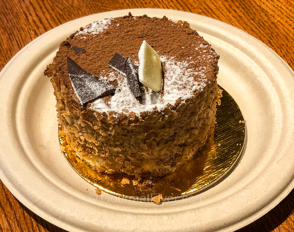 A tiramisu in a round and short cylindrical shape is served on a white paper plate.