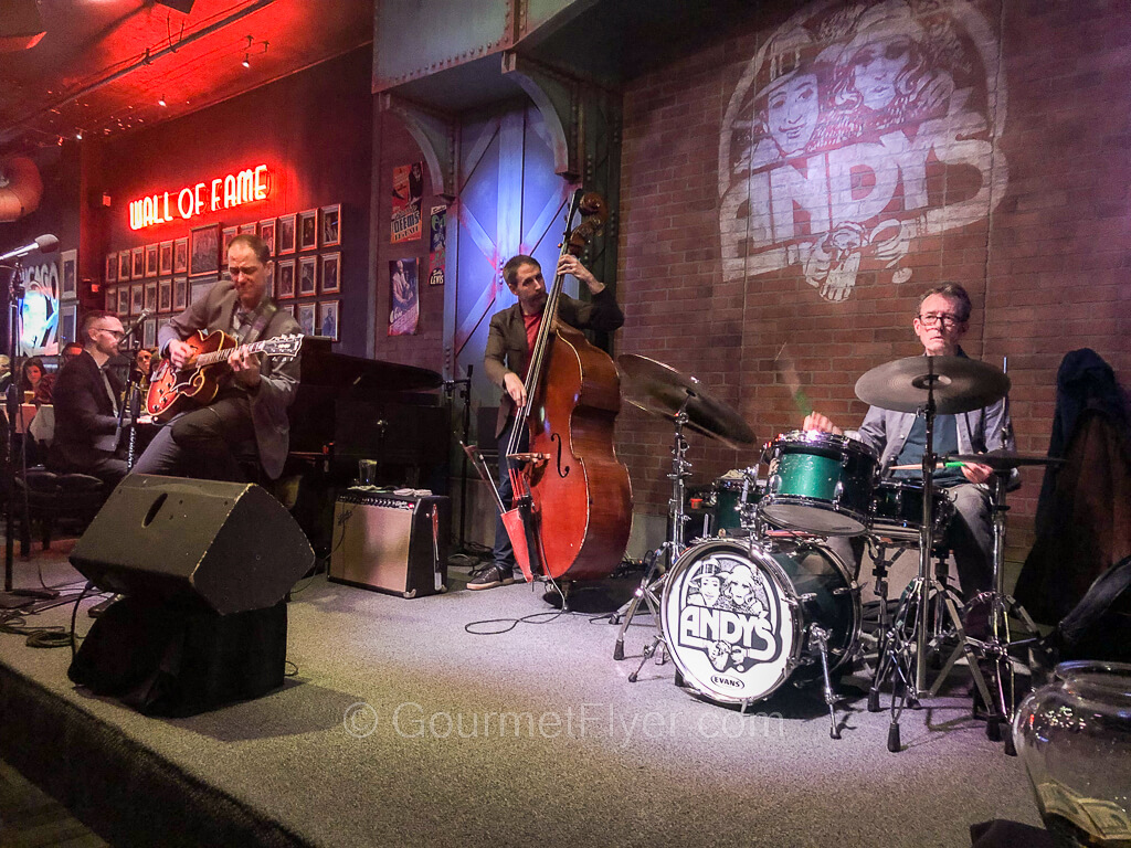 A jazz band of four men are playing on stage. From left to right, piano, guitar, bass, and drums.