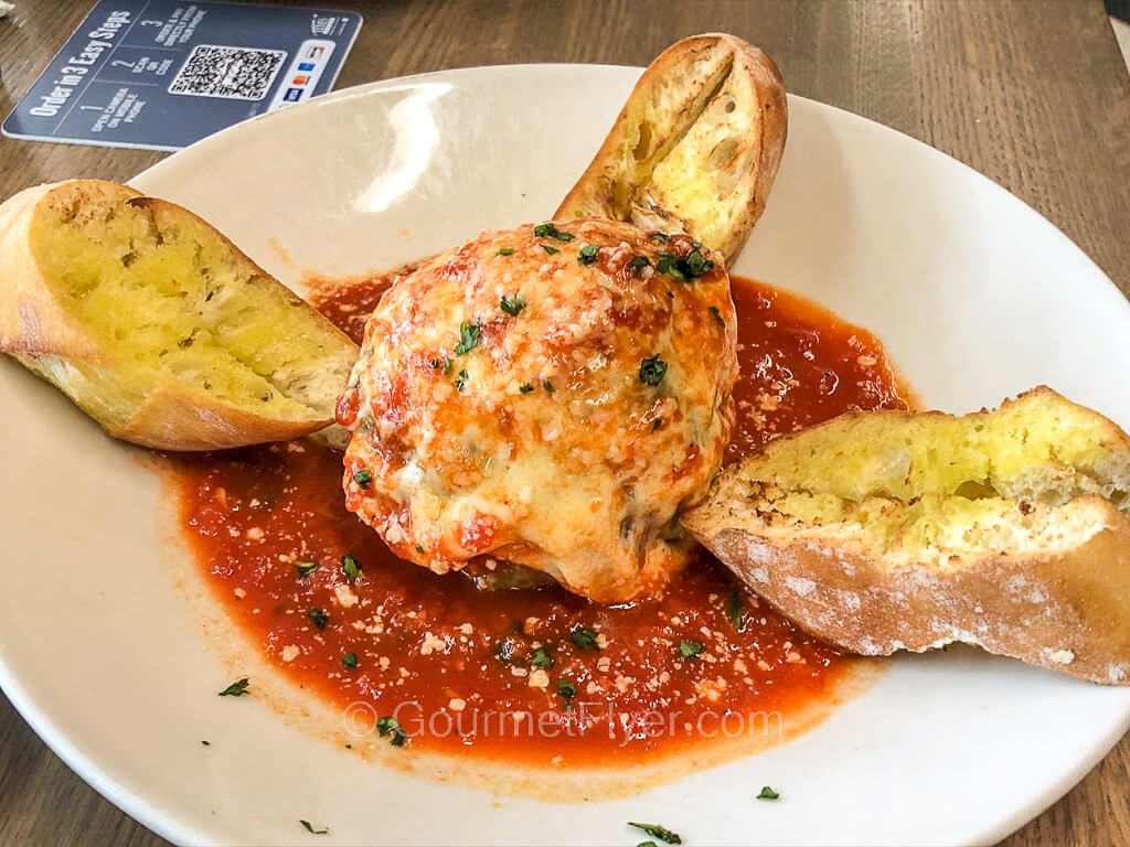 A large meatball coated with cheese is served in a dish with tomato sauce and ciabatta.