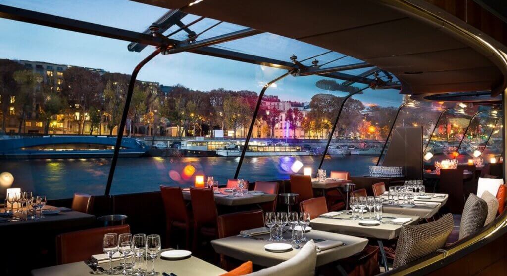 The interior of the boat with dining tables set up along the large window with night scene of Paris in the backdrop.