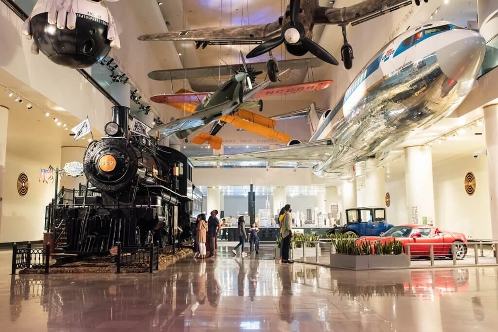 The museum hall has a train engine to the left, cars to the right, and planes hanging in the air.