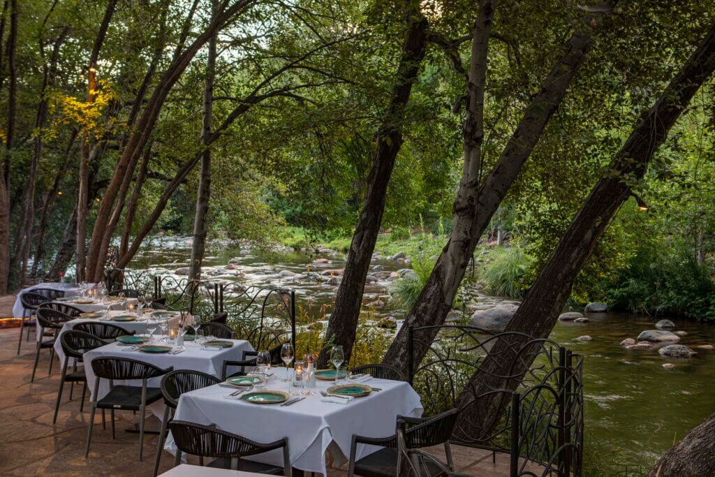 Four dining tables with white tablecloth and dinner setting line the edge of a creek.