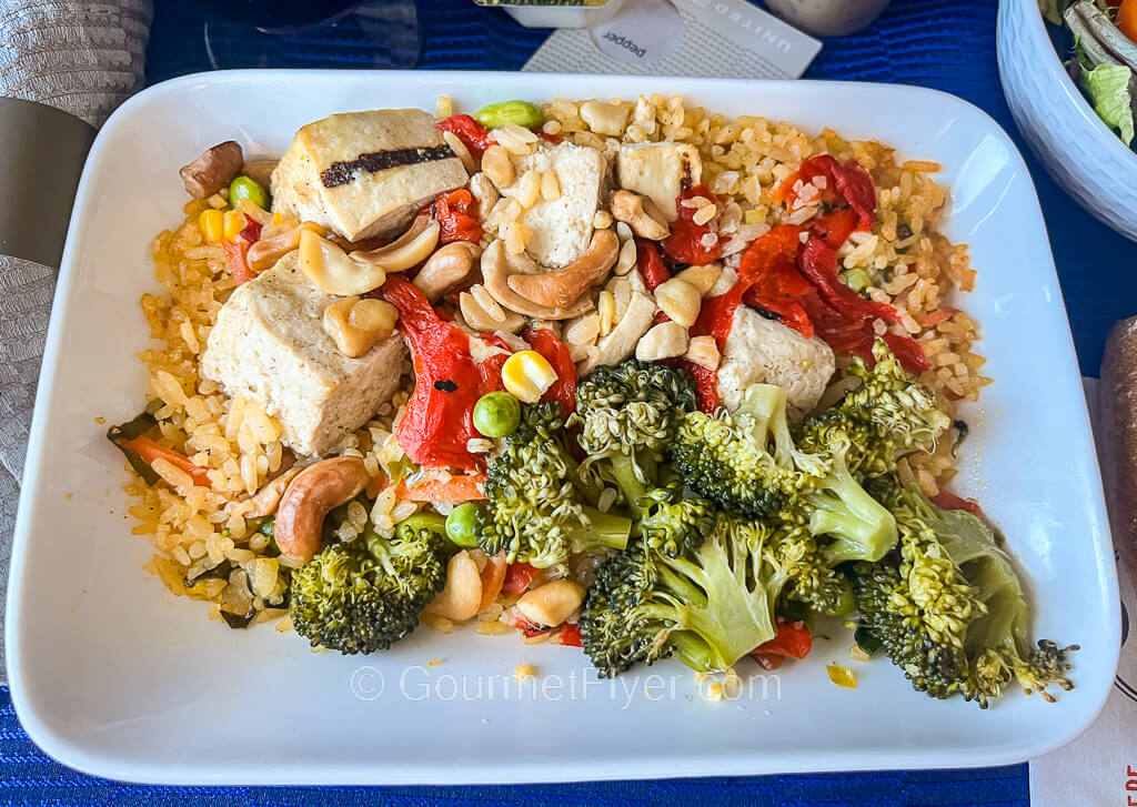 A plate of rice topped with grilled tofu, broccoli, red peppers, and nuts.