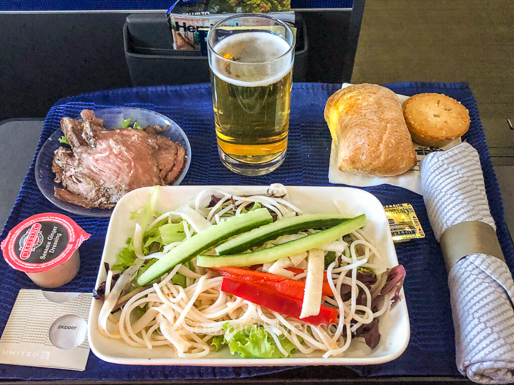An airline dinner tray consists of a glass noodle salad with beef served on the side, butter roll, dessert, and a glass of beer.