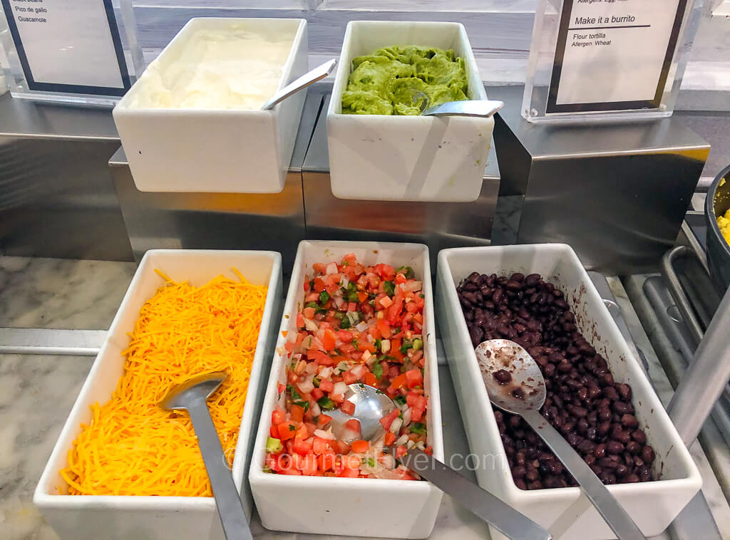 Containers of toppings such as cheese, pico de gallo and black beans are placed on a counter.