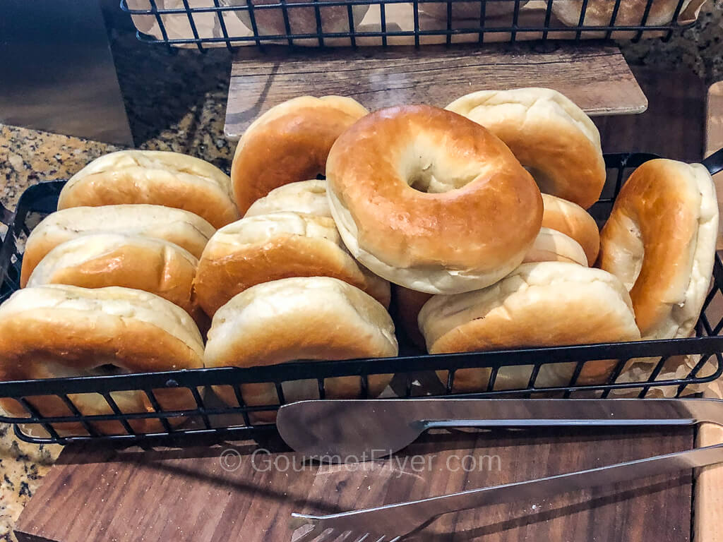 A basket of bagels is placed on a countertop with a pair of tongs.