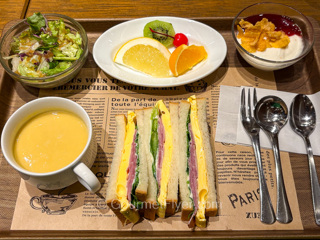 A meal tray contains a vertically presented sandwich accompanied by soup, salad, and fruits.