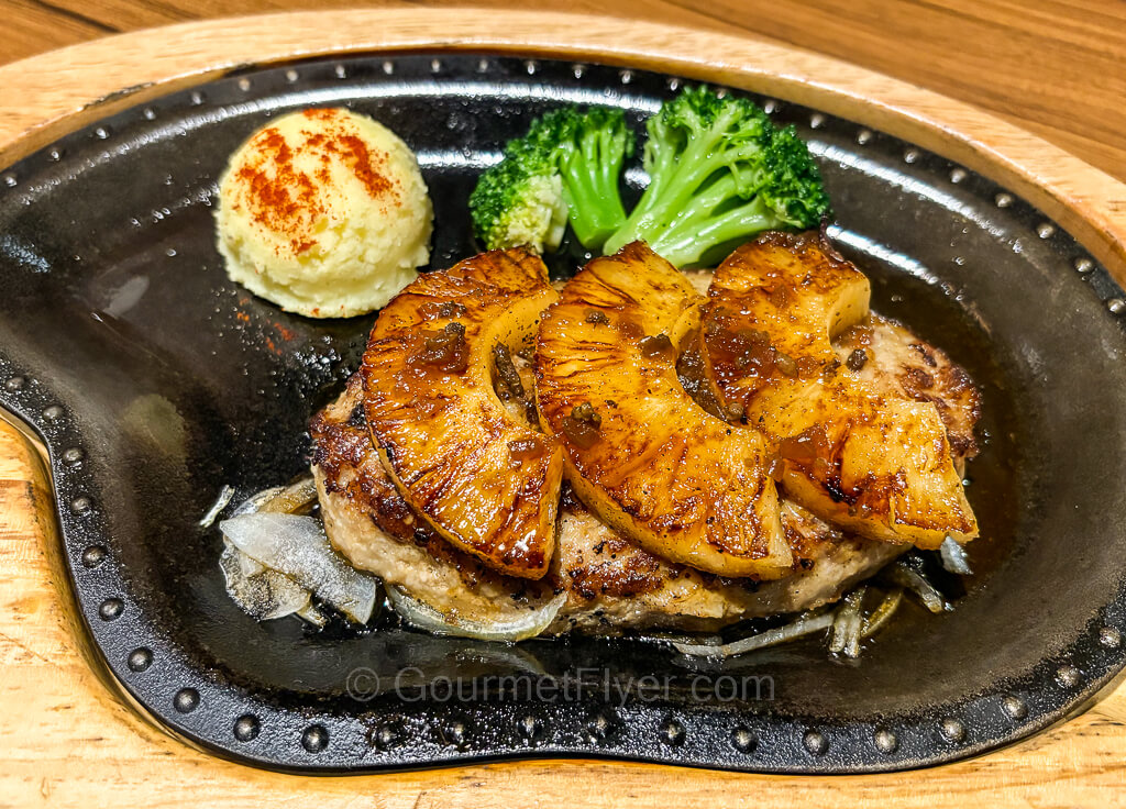 A hamburger steak topped with pineapple slices is served on a hot plate garnished with broccoli florets.