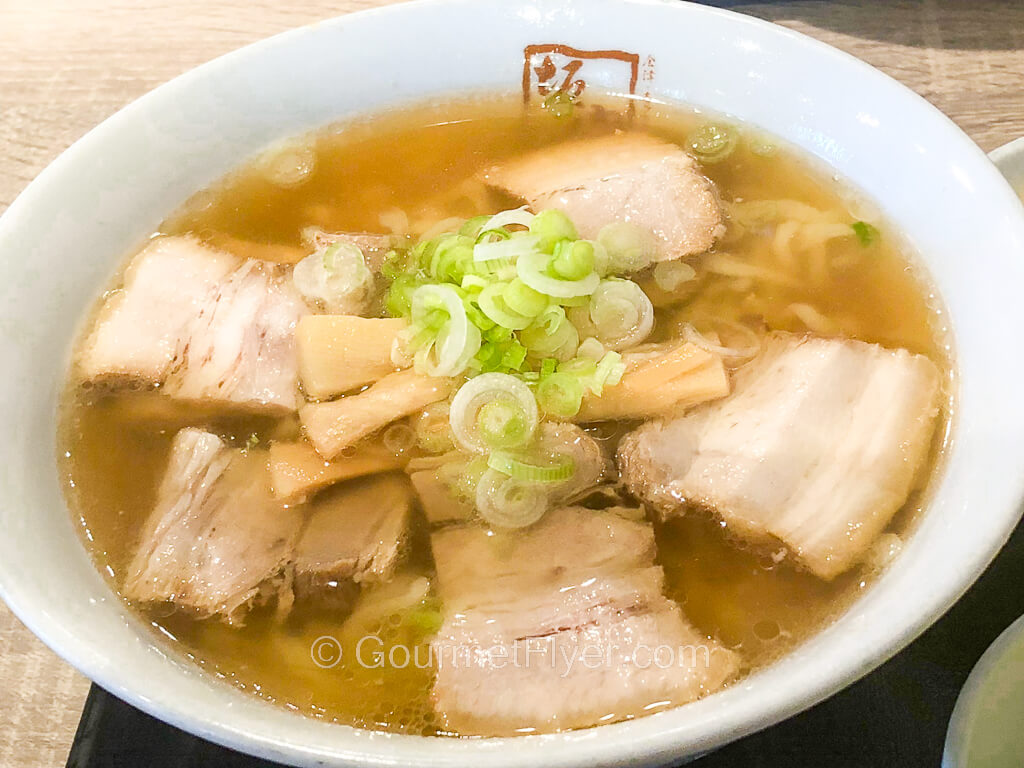 A bowl of ramen in a yellow clear broth is topped with meat and garnished with green onions.