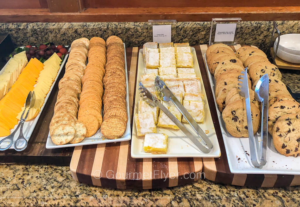 Trays of assorted cheeses, crackers, lemon cakes, and chocolate chip cookies sit atop a counter.