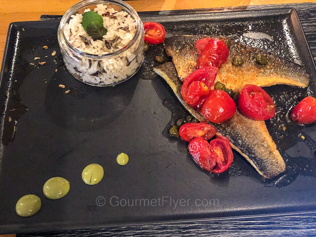 Grilled fish garnished with cherry tomatoes is served on a black platter.