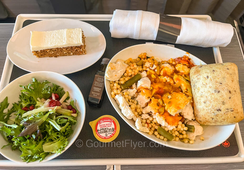 A Business Class meal tray contains a roll up with utensils, a chicken plate, side salad and a slice of carrot cake.