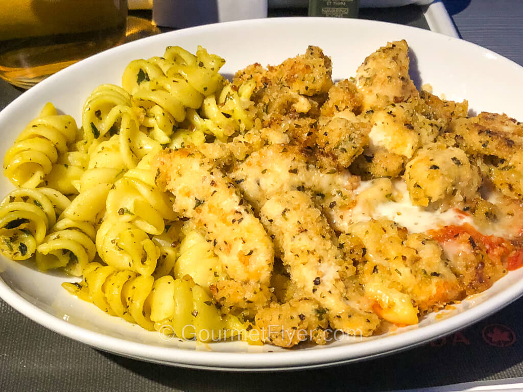 A plate of white meat coated with fried breadcrumbs is served with a side of curly pasta.