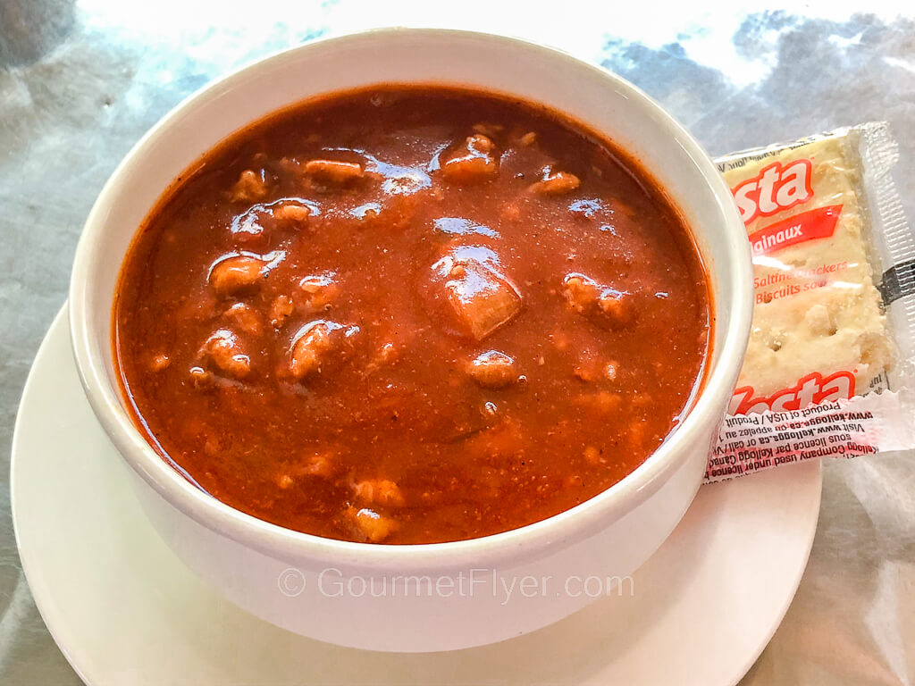 A bowl of chili sits on a plate with a package of crackers.